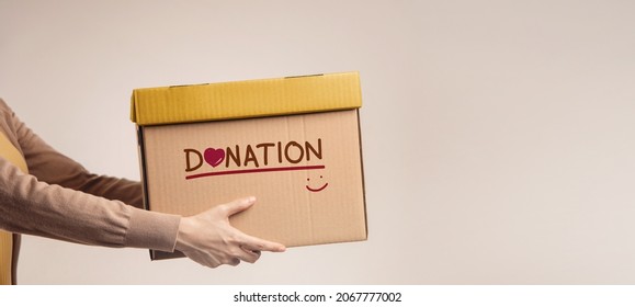 Donation Concept. Woman with Box of Things for Donate with Donation label, Smiling and Heart. Standing against the Walll. Side View