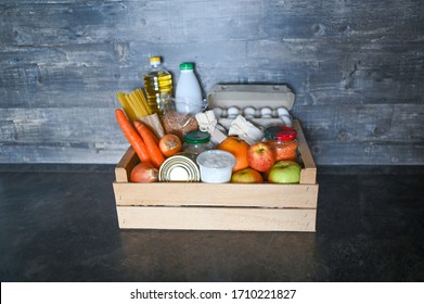 Donation Volunteer Box With Various Products For Poor Or Elderly People On Wooden Background. Pandemic Coronavirus Food Shortages. Food Donations Or Delivery Concept. Free Space For Text Copyspace