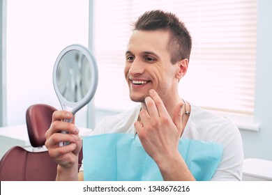 Done To Perfection. Glad Man In A Dentist Cabinet With A Mirror In His Hand And A Special Napkin On His Neck Is Smiling And Looking Through The Results Of The Dentist Great Work.