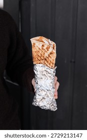 Doner kebab wrapped in flatbread held in hands, showing layers of sliced meat, fresh vegetables, and sauces, ready to eat, highlighting a casual, street food experience.	