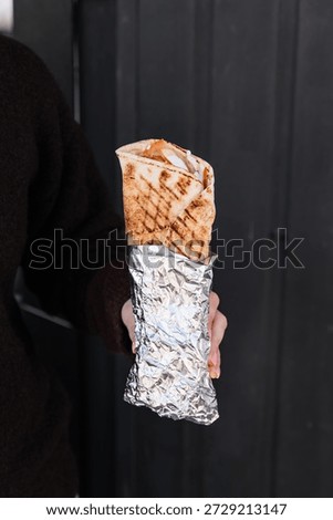 Doner kebab wrapped in flatbread held in hands, showing layers of sliced meat, fresh vegetables, and sauces, ready to eat, highlighting a casual, street food experience.	