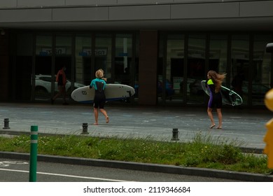 Donostia-San Sebastián, Spain. July, 2021. Two Young Girls Wearing Wetsuits And Carrying A Surfboard Run Barefoot On The Street Close To The Kursaal Convention Center And Auditorium Building.