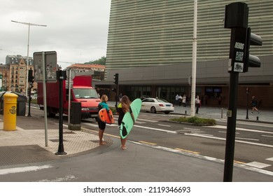 Donostia-San Sebastián, Spain. July, 2021. Two Young Girls Wearing Wetsuits And Carrying A Surfboard Wait Barefoot To Cross A Street Close To The Kursaal Convention Center And Auditorium Building.