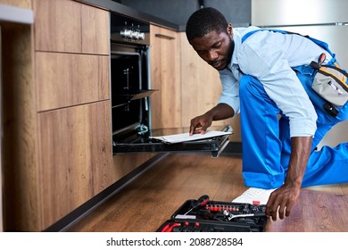 Don't delay with repair. Side view afro repairman examining oven in kitchen with tool case. Confident professional black repairman handyman in blue workwear overalls during work at home - Shutterstock ID 2088728584