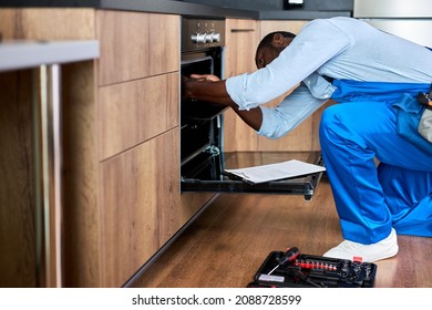 Don't delay with repair. Side view afro repairman examining oven in kitchen with tool case. Confident experienced black repairman handyman in blue workwear overalls during work at home - Shutterstock ID 2088728599