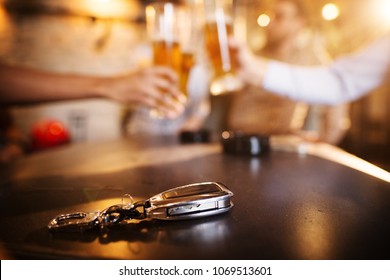 Don't drink and drive! Car key on a wooden pub table in front of blurred friend clinking with a beer.