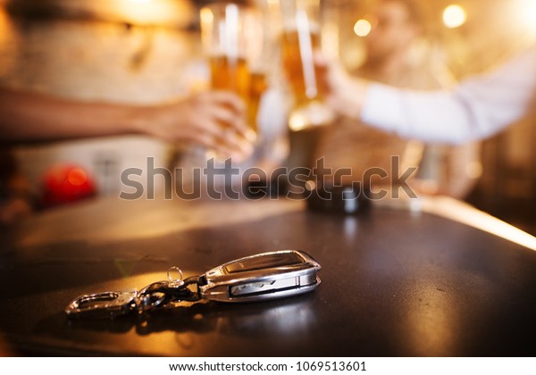 Don\'t drink and drive! Car\
key on a wooden pub table in front of blurred friend clinking with\
a beer.