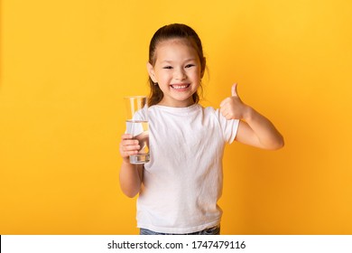 Don't Forget To Drink Water. Cute Asian Child Holding Glass Of Water And Showing Thumbs Up, Isolated Over Yellow Wall