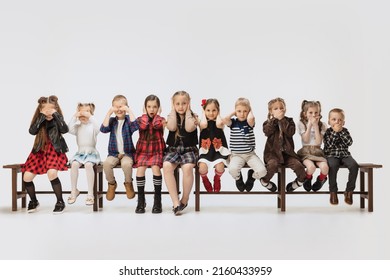 I don't hear, I don't see, I won't tell anyone. Little girls and boys, funny children sitting on banch isolated on grey studio background. Beauty, kids fashion, education, happy childhood concept.