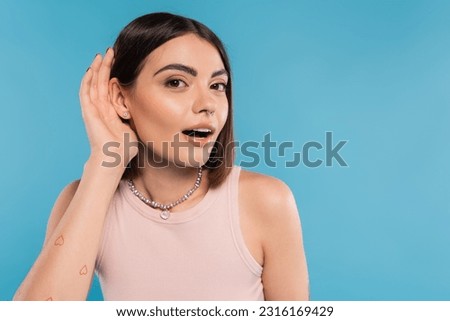 Don't hear you gesture, tattooed young woman with nose piercing and short hair holding hand near ear on blue background, generation z, listening, opened mouth, curious