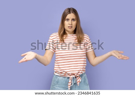 I don't know. Blond woman wearing striped T-shirt standing with wide raised arms and looking at camera with frowning face, and dont know what to do. Indoor studio shot isolated on purple background.