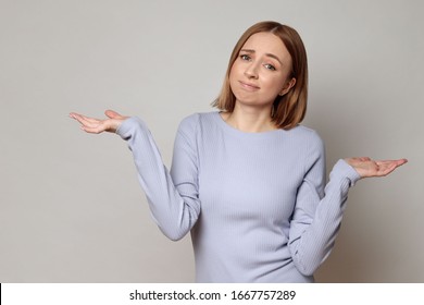 I Don't Know! Close Up Studio Portrait Of Shy Awkward Young Woman Feeling Embarrassed, Confused And Puzzled, Shrugs Shoulders, Does Not Know What Happened, Over Grey Background.