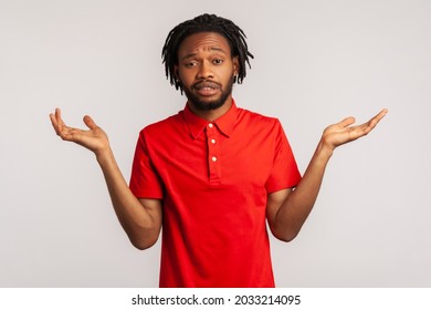 I don't know! Handsome bearded man with dreadlocks wearing red casual style T-shirt, standing with raised arms and looking at camera with answer. Indoor studio shot isolated on gray background.
