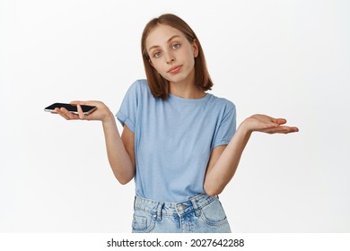 I Dont Know, Not Even Care. Young Woman Shrugging While Holding Mobile Phone, Looking Reluctant And Careless At Camera, Nothing To Say, Standing Over White Background