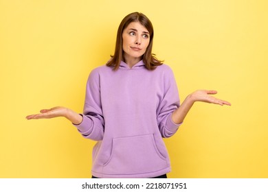 I Don't Know. Portrait Of Confused Beautiful Young Woman Standing With Raised Arms And Looking At Camera, Wearing Purple Hoodie. Indoor Studio Shot Isolated On Yellow Background.