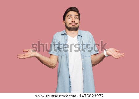 I don't know. Portrait of confused handsome bearded young man in blue casual style shirt standing with raised arms and looking at camera with answer. indoor studio shot, isolated on pink background.