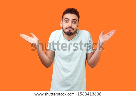 I don't know, who cares. Portrait of confused clueless brunette man with beard in white t-shirt shrugging shoulders, making no idea gesture, whatever. indoor studio shot isolated on orange background