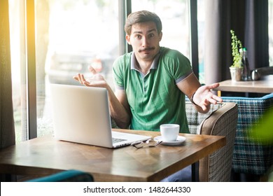 I don't know! Young confused businessman in green t-shirt sitting with laptop, looking at camera with raised arms and puzzled. business and freelancing concept. indoor shot near big window at daytime.