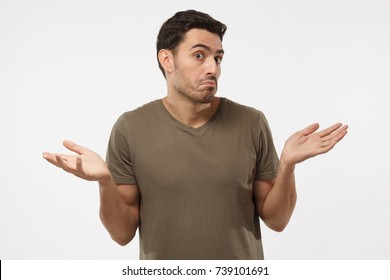 I don't know. Young man isolated on grey background being at a loss, showing helpless gesture with arm and hands, mouth curved as if he does not know what to do