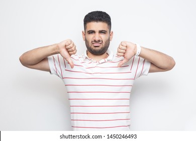 I don't like this. Portrait of displeased bearded young man in striped t-shirt standing with thumbs down dislike sign gesture and looking at camera. indoor studio shot, isolated on white background.