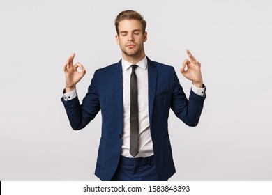 Dont panic, taking emotions under control. Peaceful and patient good-looking blond businessman in classic suit, make zen sign, spread hands sideways, close eyes, meditating, reliese stress