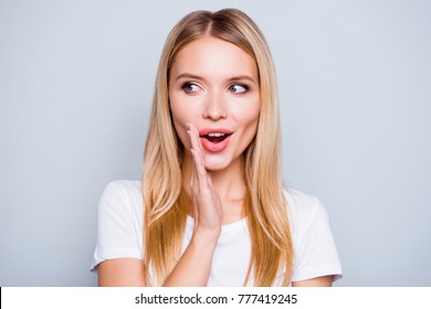 Don't tell anyone! Talkative mysterious pretty beautiful woman with blonde hair dressed in white tshirt is saying secret hot braking news and looking aside, isolated on grey background