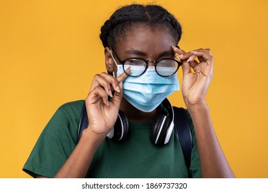 Don't Touch Your Face. Black Female Student Wearing Medical Mask And Scratching Eye Standing Over Yellow Studio Background. Covid-19 Pandemic Outbreak, Coronavirus Protection