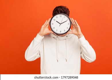 Don't Waist Your Time! Man In Casual White Sweatshirt Holding Wall Clock Hiding His Face, Time Management, Schedule And Meeting Appointment. Indoor Studio Shot Isolated On Orange Background