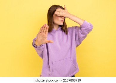 Don't Want To Look. Portrait Of Woman Keeping Hand On Eyes And Avoiding Watch At Something Shameful, Showing Stop Ban Gesture, Wearing Purple Hoodie. Indoor Studio Shot Isolated On Yellow Background.