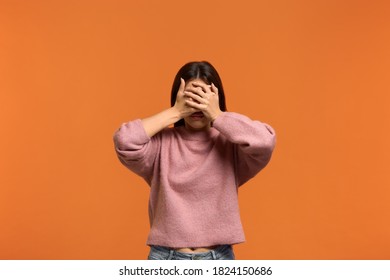 I dont want to see this. Portrait of woman in pink sweater, makes denial hand gesture, demonstrates stop sign by palm, and covers eyes with palm says no to somebody. isolated on orange wall