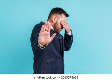 I dont want to see this. Side view portrait of man wearing official style suit standing, closed his eyes with hands and showing stop gesture. Indoor studio shot isolated on blue background.