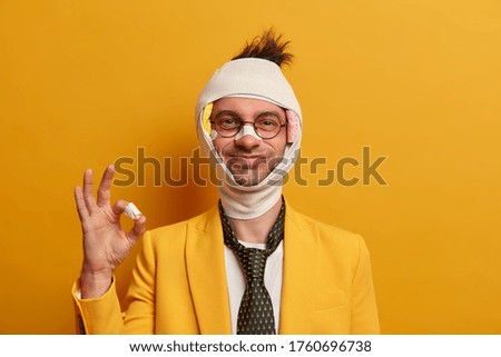 Dont worry, everything is okay. Smiling positive adult man forgets about accident consequences and injuries, gestures ok sign, received first aid from doctor, wears elegant suit, bandage on head