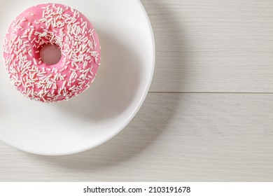 Donut With Pink Glaze, Lying On White Plate On Light Wood Countertop. Top View.