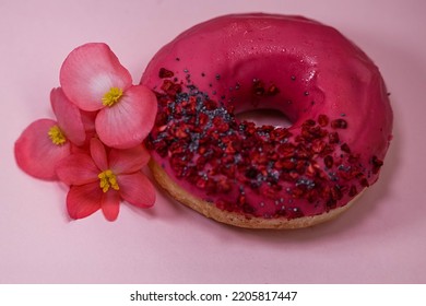 Donut With Pink Icing And Colored Sprinkles, Poppy Seeds And Dry Raspberry On Pink Background With Edible Rose Begonia Petals Flowers. Tasty Delicious Fat High Calories Sweet Food. Selective Focus