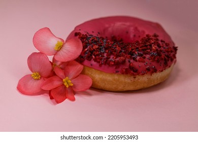 Donut With Pink Icing And Colored Sprinkles, Poppy Seeds And Dry Raspberry On Pink Background With Edible Rose Begonia Petals Flowers. Tasty Delicious Fat High Calories Sweet Food. Selective Focus