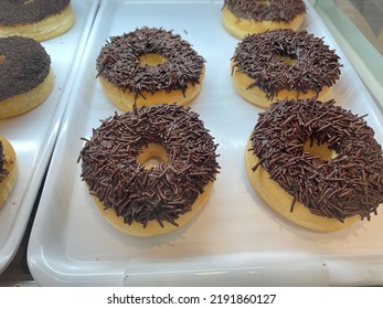 Donuts With Chocolate Sprinkles Lined Up On A White Tray In A Glass Display Case