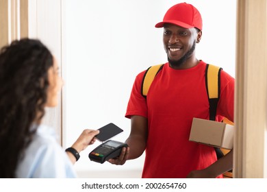 Door Delivery. Back View Of Customer Lady Receiving And Paying For Package Box With Phone. Smiling Black Courier Guy In Red Uniform Standing In Doorway At Home Holding Parcel. Modern Shopping Concept