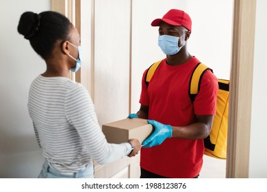 Door Delivery. Black Courier Wearing Protective Face Mask And Gloves Giving Package Box To Lady Standing At Doors Of Her House. Safe Shopping, Woman Receiving Parcel During Quarantine Lockdown