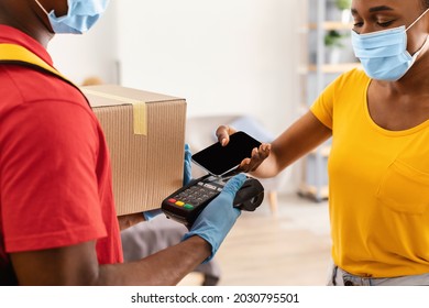 Door Delivery. Black Woman Receiving Package Box From Deliveryman And Paying With Smartphone For Courier Service At Home, Wearing Protective Face Mask. Selective Focus, Cropped