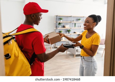Door Delivery. Happy Black Customer Lady Receiving And Paying For Package Box With Phone Taking Parcel From Courier Guy In Red Uniform Standing In Doorway At Home. Modern Shopping Concept