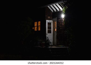 Door To A House At Night. Golden Light Shining From Window, Porch Light On.   Cedar Shake Shingles On House. Green Trees To Sides Of Door.