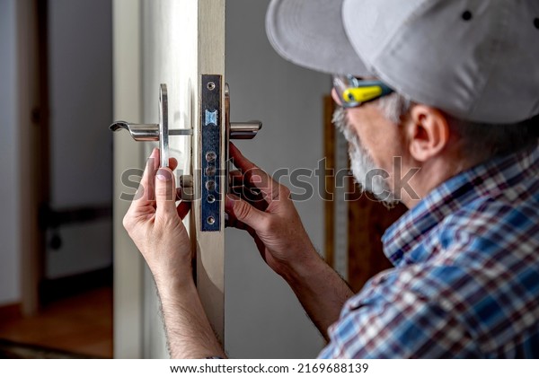 Door lock installation, repair, or replacement\
service. Door hardware installer locksmith working with open white\
door indoor.