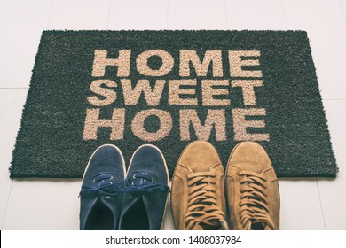 Door Mat Shoes At Front Entrance Of Condo Apartment. Written Welcome Sign Home Sweet Home Welcoming Homeowners At New House Moving In Couple's Pairs Of Sneakers Lying On The Floor.