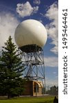 Doppler weather radar ball against blue sky with white clouds and sun reflecting off of sphere. Located at a National Weather Service site. Tree in foreground.