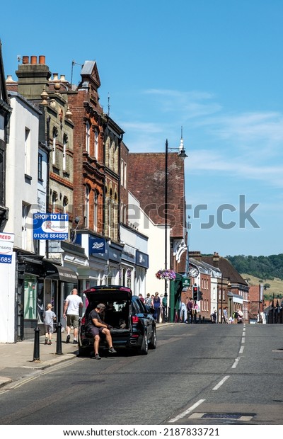 Dorking, Surrey Hills, London UK, July 07 2022, Row\
Of Retail Shops Dorking High Street Against A Blue Sky With people\
Walking And Parked Car