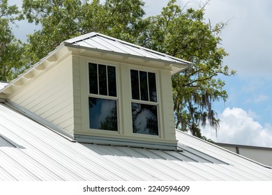 A dormer with multiple double hung windows, a grey metal roof, and pale yellow vinyl siding on a large house. There are tall trees with thick green leaves both in front and behind the building. 