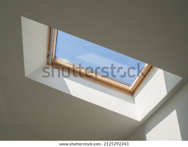 Dormer wooden window in the white sloping ceiling\
overlooking the blue sky. Sunlight enters the room through a closed\
window