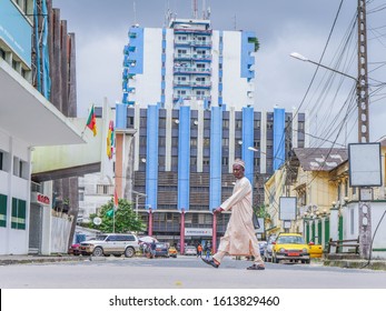 Douala / Cameroon - January 12 Th 2020. A Man Crossing The Street, With A Blue And White Building Behind With Cameroon Flag In The Very Heart Of Douala, 