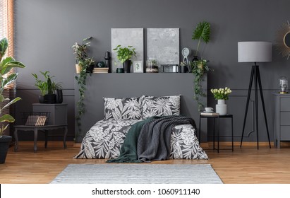 Double Bed With Floral Bedding And Dark Blankets Standing In Grey Bedroom Interior With Fresh Plants, Paintings And Lamp