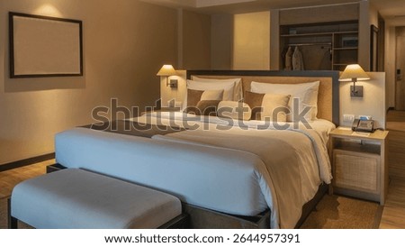 A double bed with white sheets and a beige bedspread. Pillows at the headboard. The couch. The phone is on the bedside table. The lamps are lit. The interior of a bedroom in a hotel. The suite.